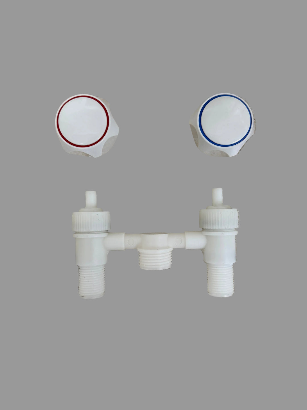 White plastic dual-handle valve trim set with two white hexagonal knobs, one featuring a red ring and the other a blue ring, positioned above a matching horizontal cross-connecting pipe assembly on a speckled beige stone surface.